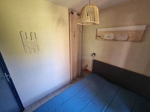 A bedroom with a bed, a hanging light fixture, a wall-mounted shelf with decorative items, and a door with a towel rack.