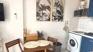 Une cuisine compacte avec une table en bois, des chaises, un four à micro-ondes, un lave-linge et deux reproductions botaniques encadrées accrochées au mur.