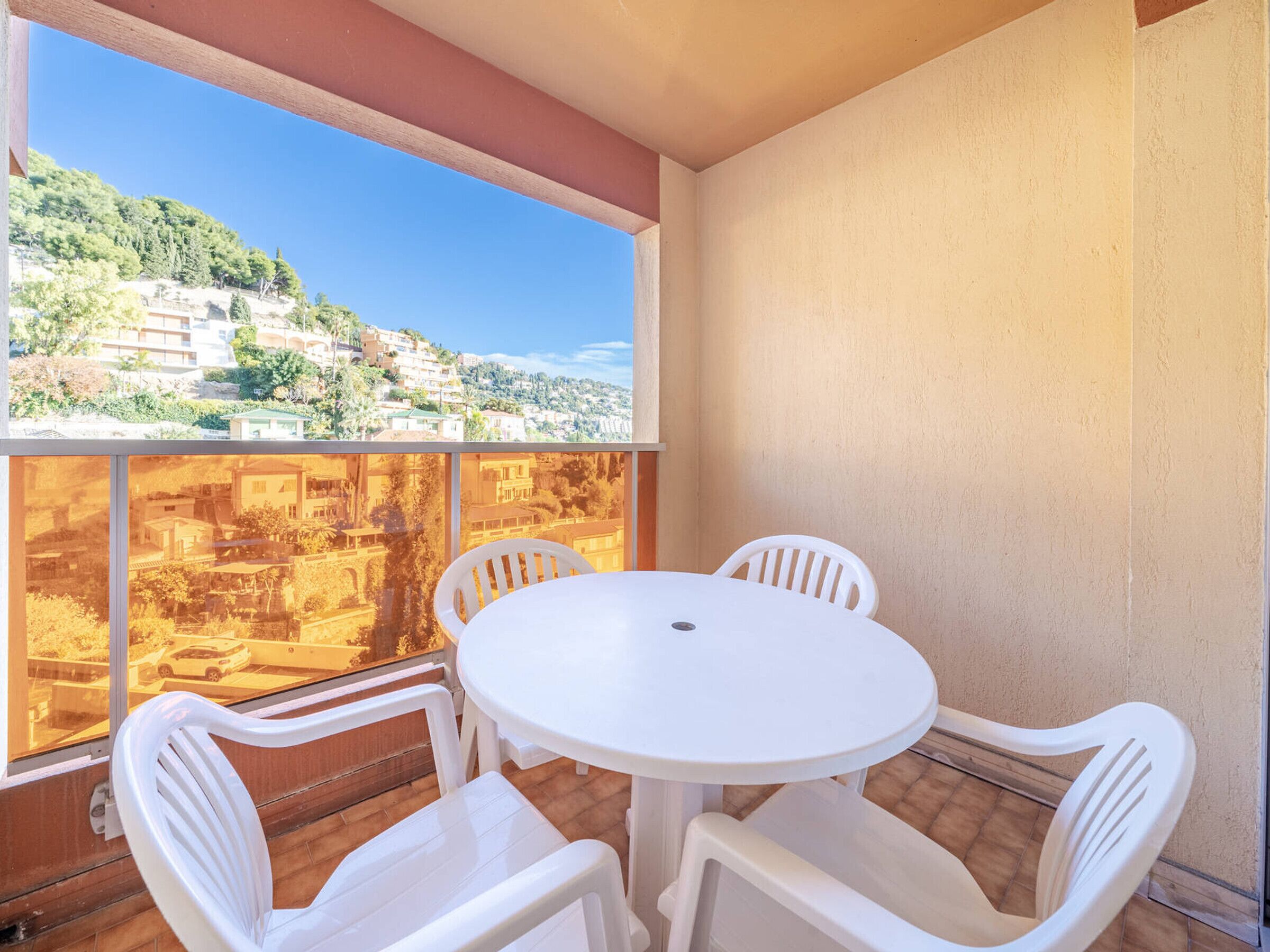 A balcony with a white table and four chairs, overlooking a hillside with buildings.