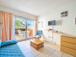 A modern living room with a sofa, a blue armchair, a wooden coffee table, a TV mounted on the wall, and a balcony with a view of buildings and greenery. - Studio for 4 with WiFi, sea view (Roquebrune-Cap-Martin)