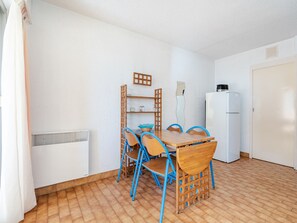 A small, clean room with a wooden dining table, blue chairs, a white refrigerator, and a tiled floor.