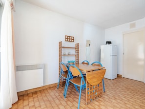 A small, clean room with a wooden dining table, blue chairs, a white refrigerator, and a tiled floor.