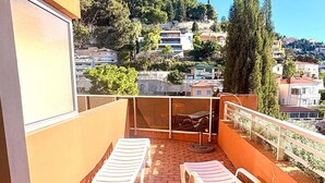 Ein Balkon mit zwei Liegestühlen und einem Tisch, mit Blick auf eine Hügellandschaft mit Gebäuden.