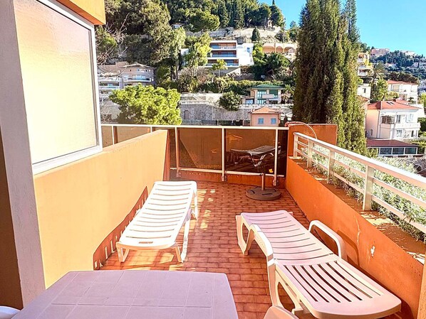 A balcony with two sun loungers and a table, overlooking a hillside with buildings.