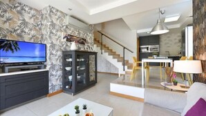 A modern living room with a flat-screen TV, a dining area with a white table, and a kitchen in the background.