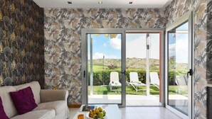 A modern living room with a sofa, a coffee table with fruits, and a sliding glass door leading to an outdoor area with chairs.