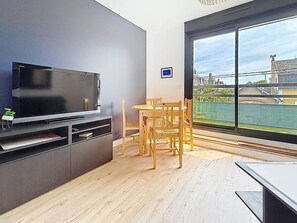 A modern living room with a flat-screen TV, wooden dining table, and chairs, a small bookshelf, and a large window with a view of buildings and greenery.