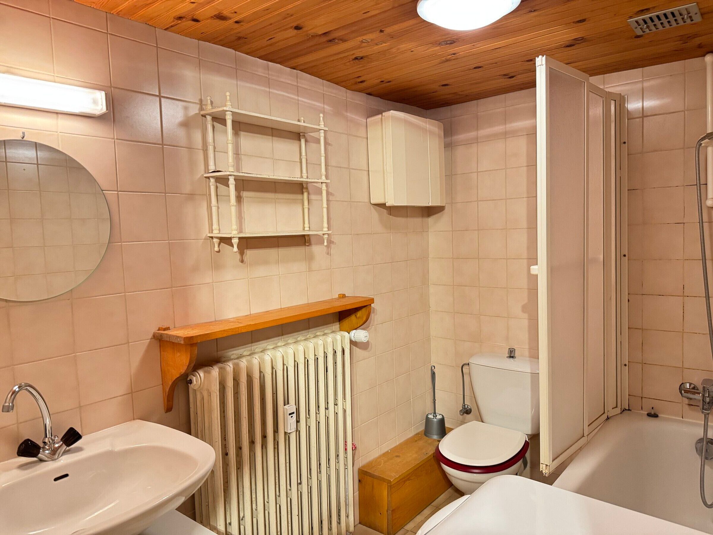 Een badkamer met een wastafel, toilet en bad. Daarboven hangt een houten plank bij de wastafel en eronder staat een radiator.