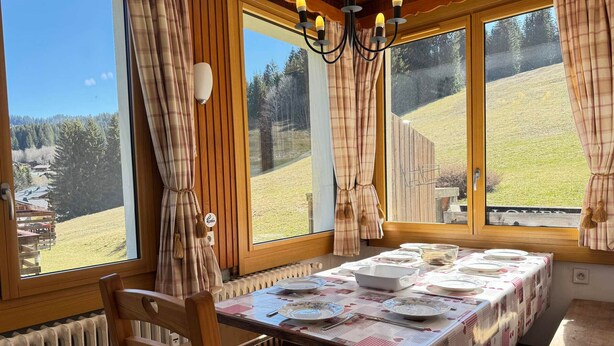 A dining area with a table set for four, large windows with curtains, and a view of a hillside.