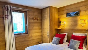 A wooden cabin room with a bed, a window with curtains, a small stool, and a wall-mounted light fixture.
