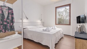 A bedroom with a bed, a hanging garment, a bedside table, a lamp, a TV, and a window with a view of trees. - Villa Toschi beach front (TERTENIA)