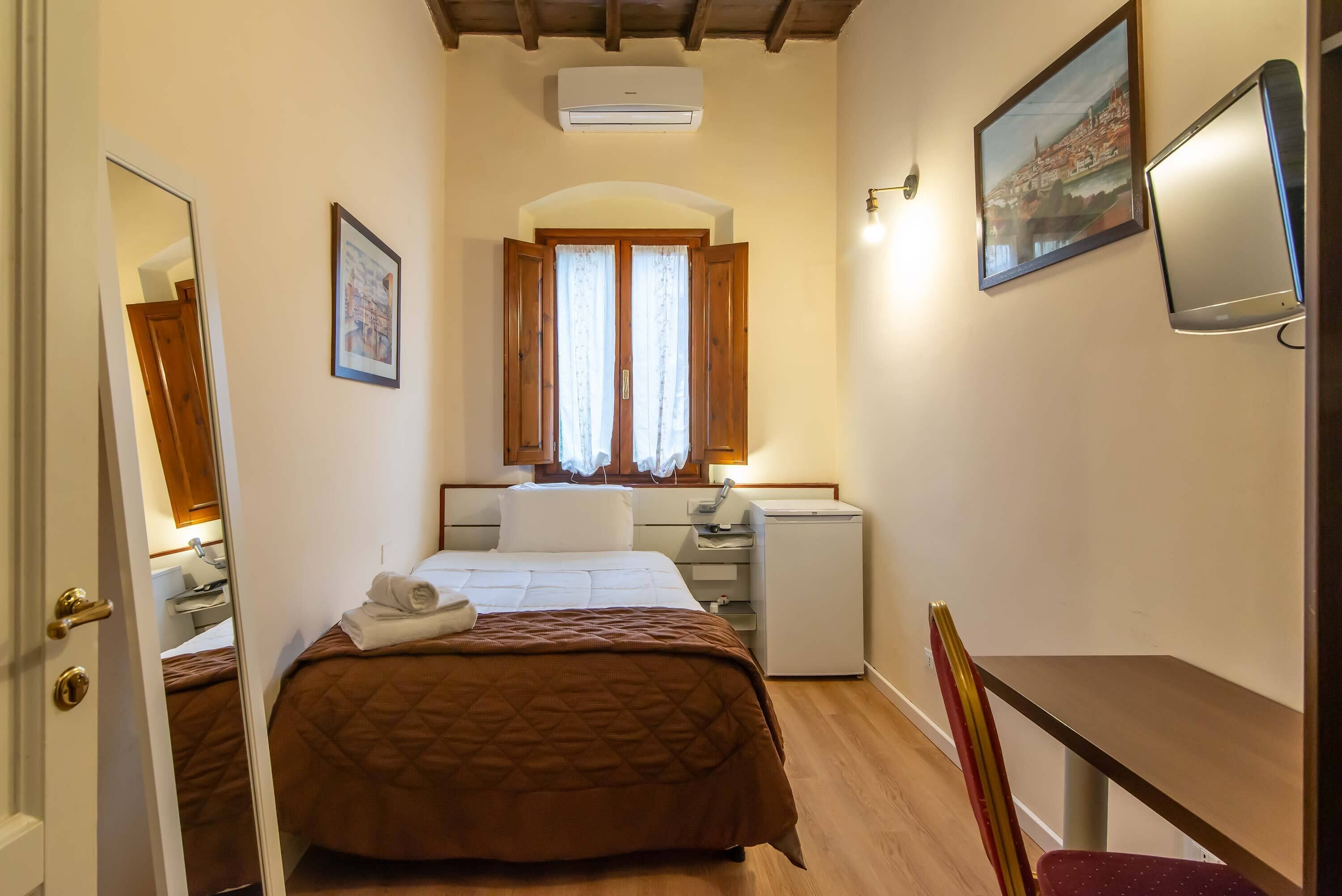 Een hotelkamer met een bed, een bureau, een stoel, een bureaulamp, een kleine koelkast en een raam met gordijnen.