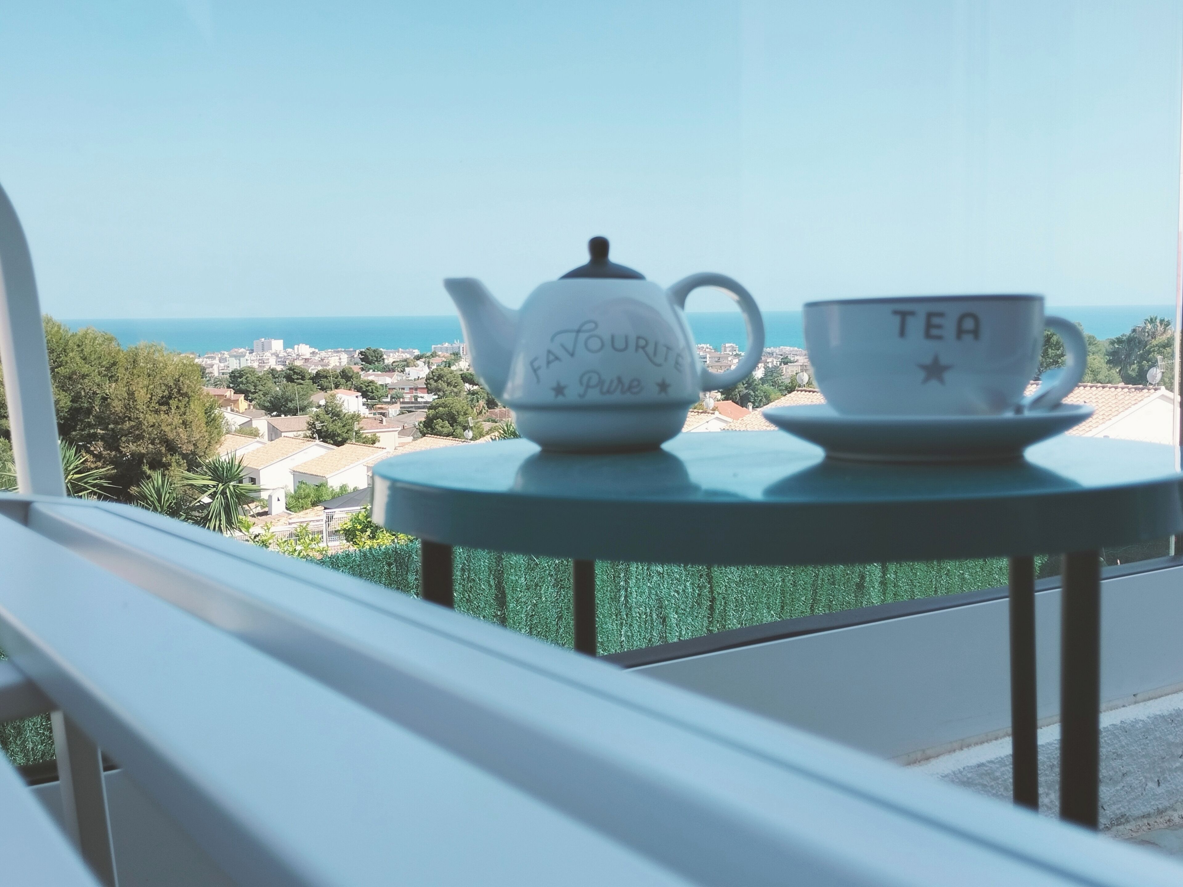 A balcony with a table holding a teapot and a cup, overlooking a coastal town.