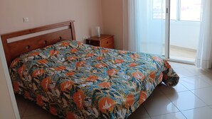 Een slaapkamer met een bed, een nachtkastje, een raam met gordijnen en uitzicht op buiten.