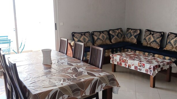 Een eetruimte met een lange tafel, overdekt met een tafelkleed met dessin, omringd door houten stoelen. Tegen de muur staat een bank met gekleurde kussens.