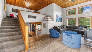 A spacious living area with a wooden staircase, a blue armchair, a sectional sofa, and a fireplace. - Black Fork Lodge - Beautiful log home on 2.5 acres with trailer parking and panoramic views (McCall)