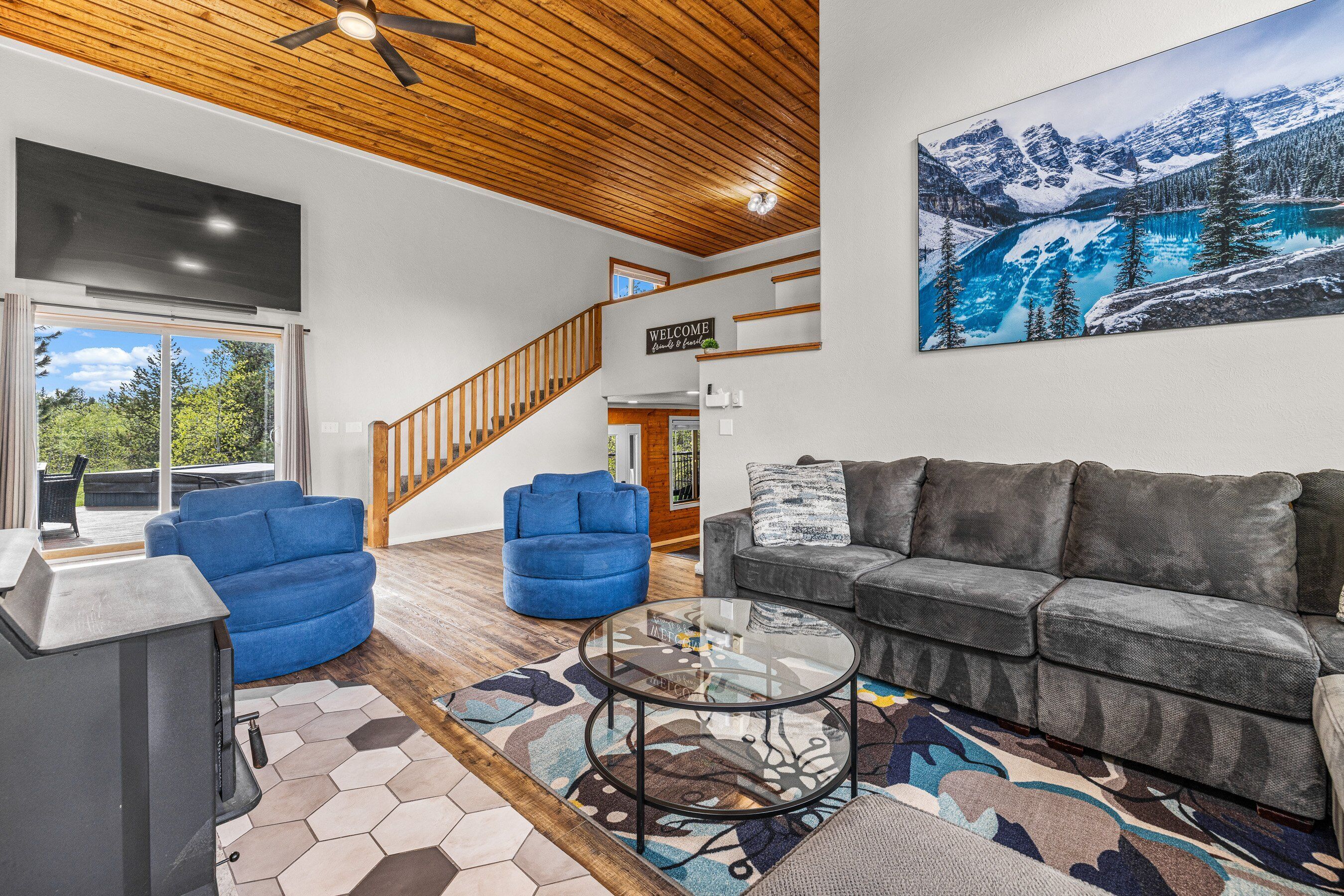 Uma sala de estar espaçosa com teto de madeira, uma grande pintura de paisagem de montanhas, um sofá cinza, poltronas azuis e uma mesa de centro de vidro.