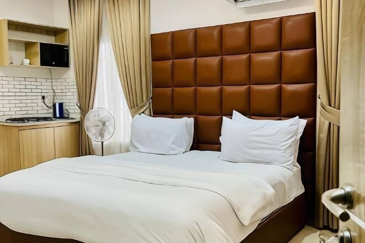 A neatly arranged hotel room with a brown leather headboard, white bedding, a small bedside table with a fan, and a shelf with a microwave and a cup.