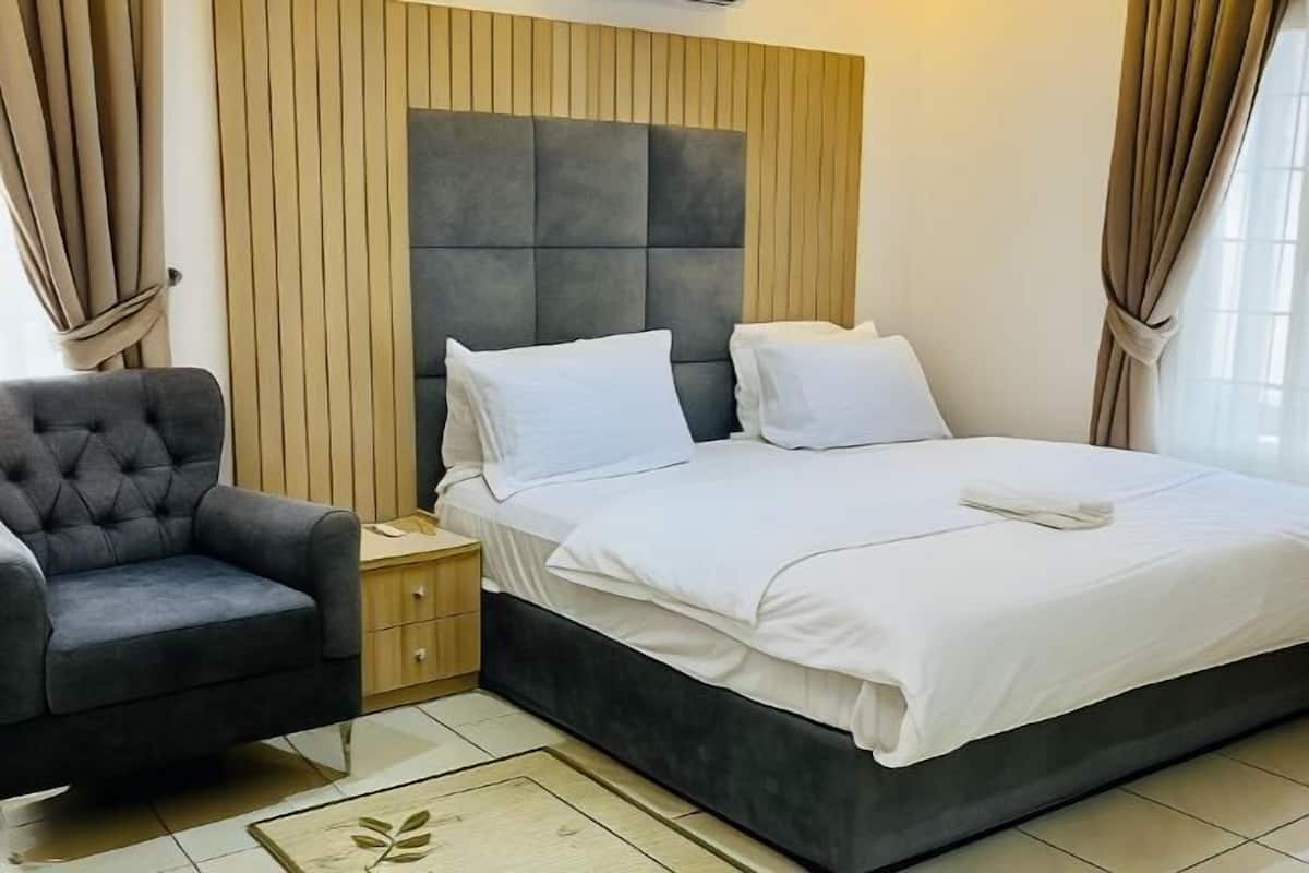 A bedroom with a bed, a nightstand, a chair, and a wooden headboard.