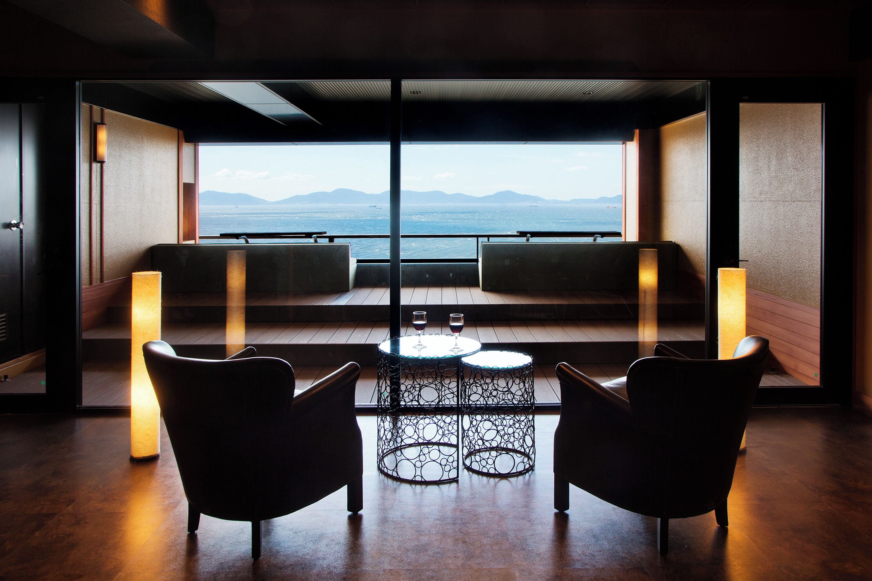 A modern room with a balcony, two chairs, a small table, and a view of the sea and mountains.