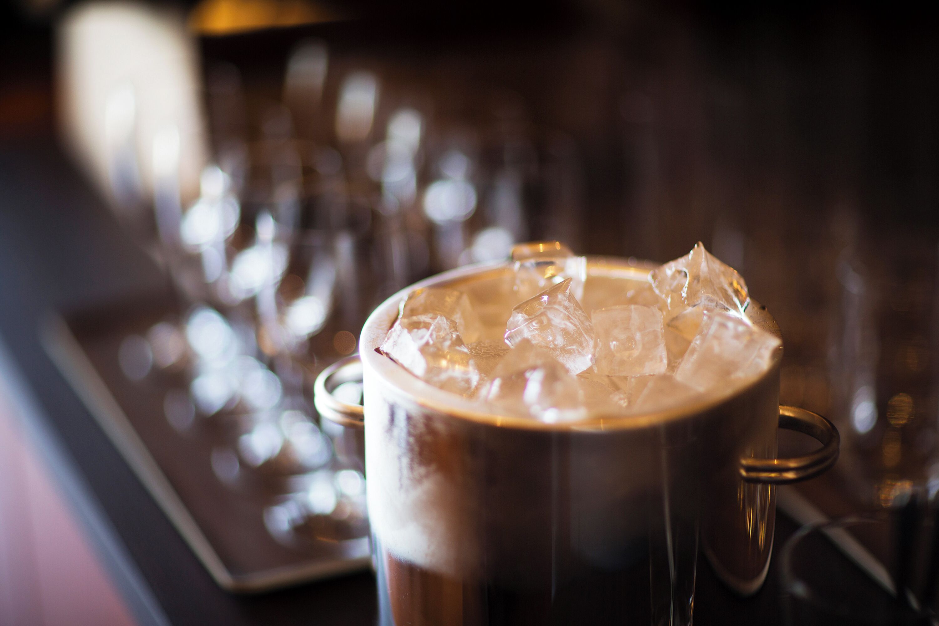 A close-up of a metallic mug filled with a dark beverage and ice cubes, with a blurred background of similar mugs.