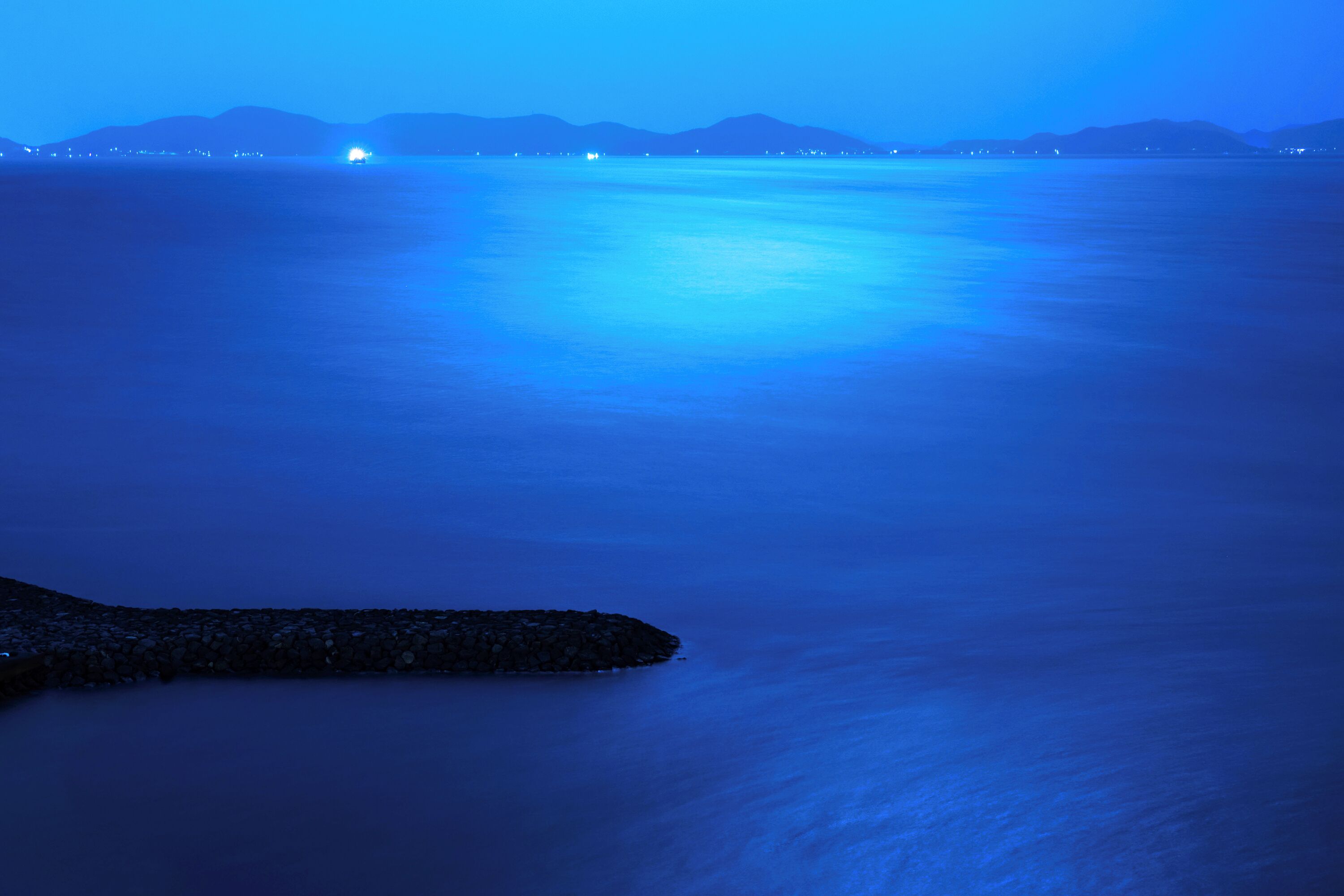 Una masa de agua tranquila con un promontorio rocoso en primer plano y montañas lejanas bajo un cielo azul.