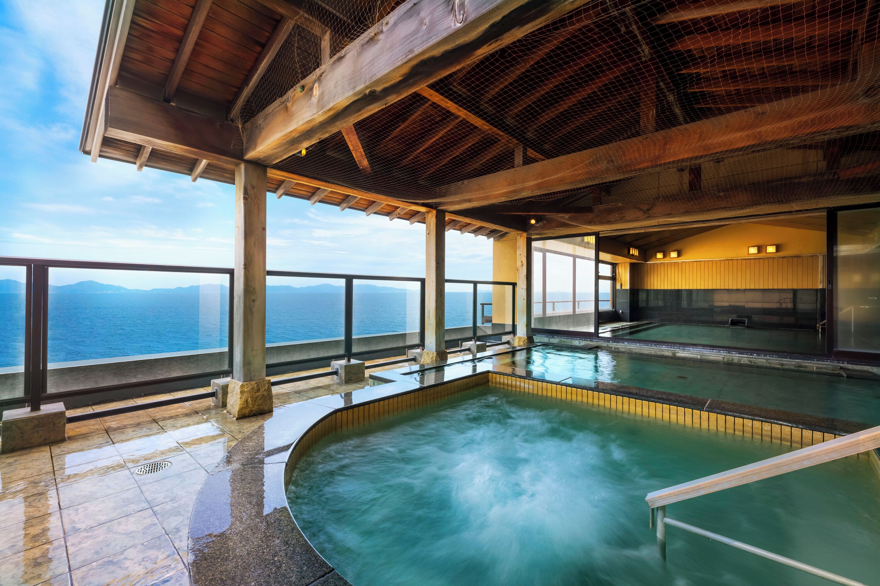 Un área de spa con hidromasaje y piscina, amplias ventanas con vistas al mar y una estructura de madera cubierta.