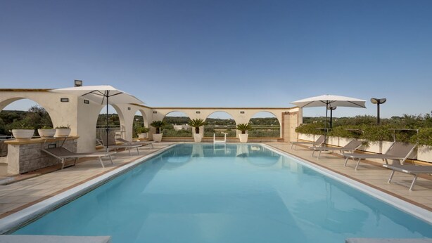 Une piscine rectangulaire à l’eau d’un bleu limpide, entourée de chaises longues et de parasols, avec une terrasse en pierre et en béton.