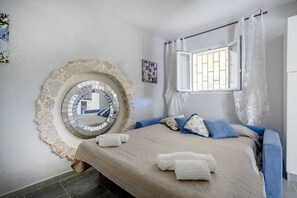 Une chambre avec un mur en pierre, un miroir rond, un lit avec une literie bleue, une fenêtre avec des rideaux et un petit tableau accroché au mur.