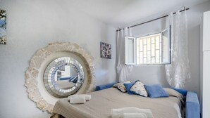 Une chambre avec un mur en pierre, un miroir rond, un lit avec une literie bleue, une fenêtre avec des rideaux et un petit tableau accroché au mur.