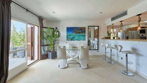 A modern dining area with a glass table and white chairs, a kitchen with bar stools, and a large window with curtains.