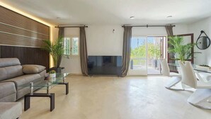A modern living room with a sofa, a glass coffee table, a flat-screen TV, and a dining area with white chairs.