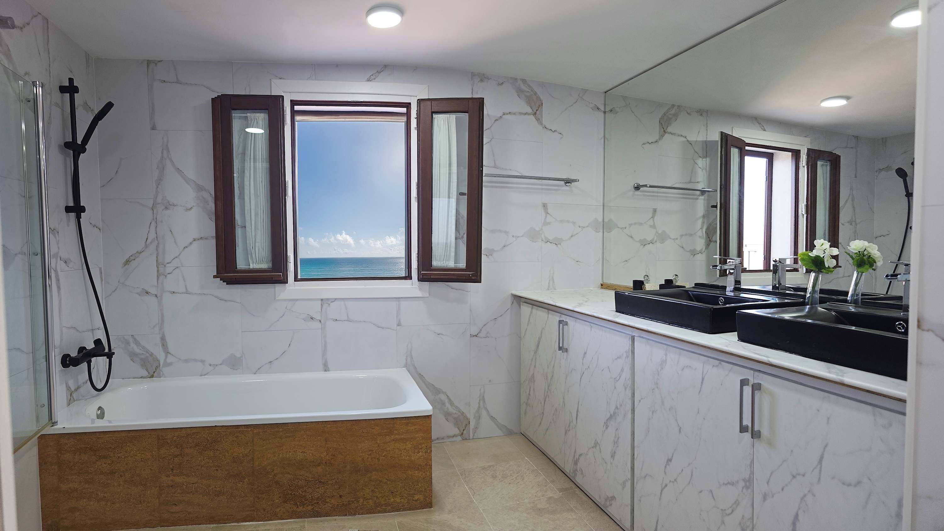 A bathroom with a bathtub, a large mirror, and a window with a view of the sea.