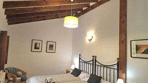 A bedroom with a bed, wooden beams, a hanging light fixture, framed pictures on the wall, and a chair.