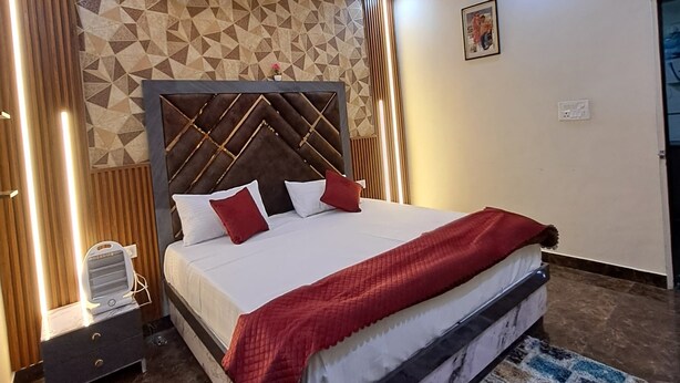 A modern bedroom with a geometric-patterned wall, a tufted headboard, a ceiling fan, and a red throw blanket. - Prem Aashiyana Home Stay (Agra)