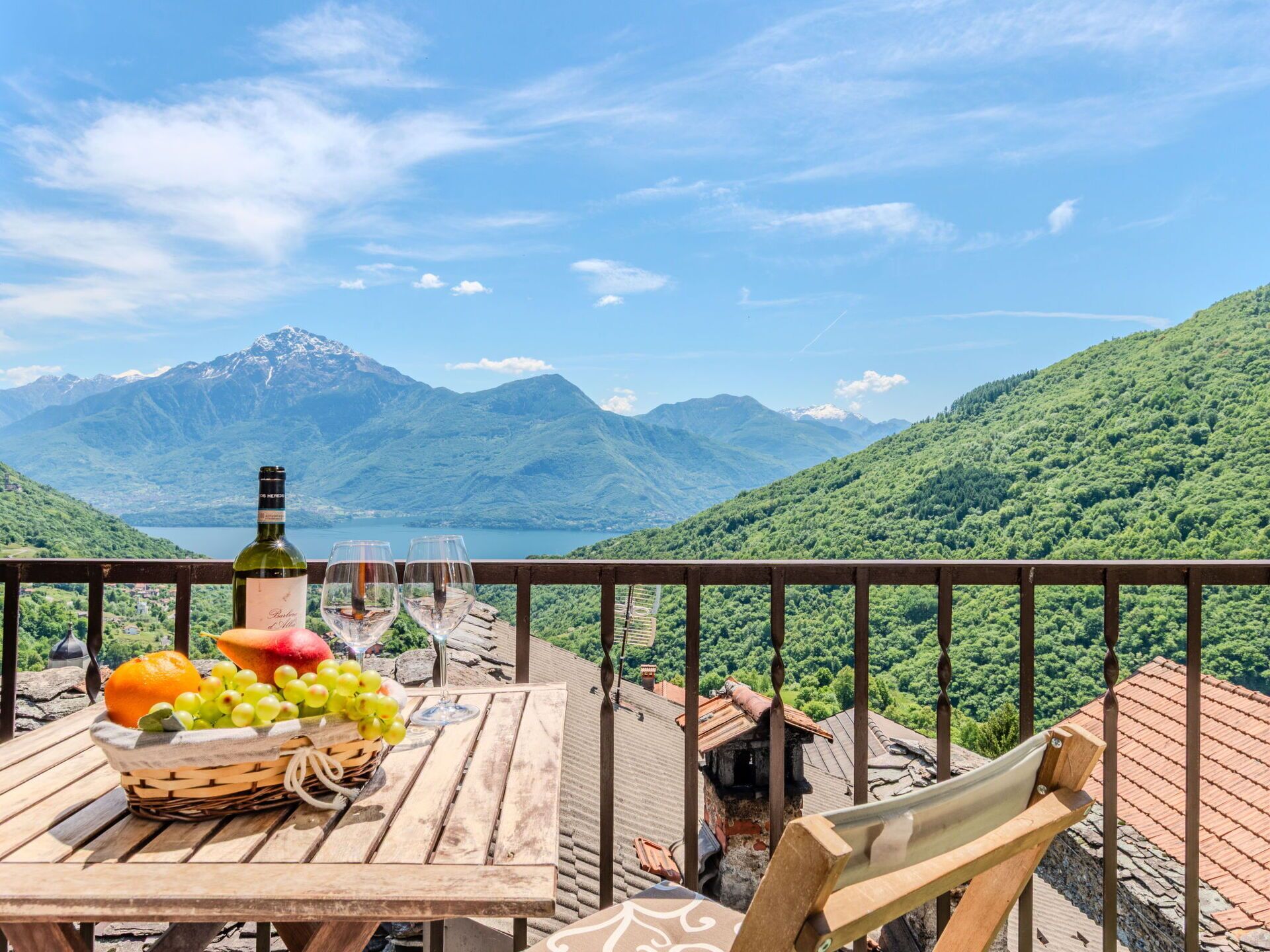 A wooden table with a bottle of wine, two glasses, and a basket of fruit on a balcony overlooking a mountainous landscape.