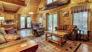 A spacious living room with a high ceiling, wooden walls, a large window, and a cozy fireplace. - Hidden Mountain Dream, Lake View, Foosball, Hot Tub, Free Attraction Tickets!!! (Sevierville)