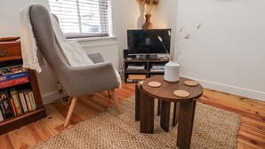 A living room with a grey armchair, a round wooden table with three coasters, a bookshelf with books, a flat-screen TV, and a vase with dried plants.