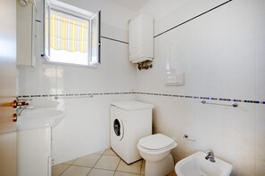 Een kleine, schone badkamer met een wastafel, toilet, wasmachine en een raam met een gestreept zonnescherm.