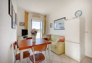 A dining area with a table and chairs, a television, a sofa, a refrigerator, and a clock on the wall.