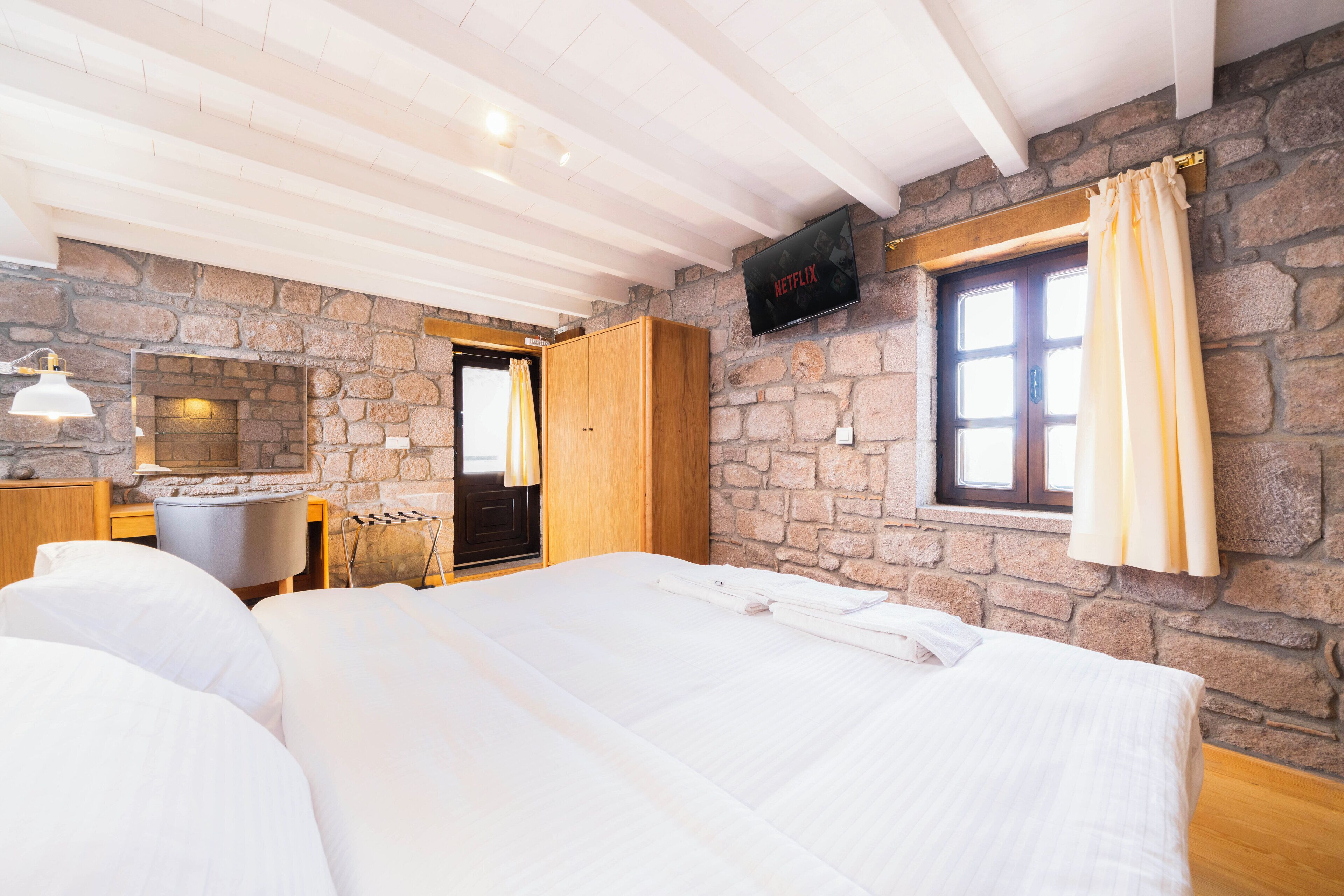 A bedroom with a stone wall, a wooden bed, a TV mounted on the wall, and a window with curtains.