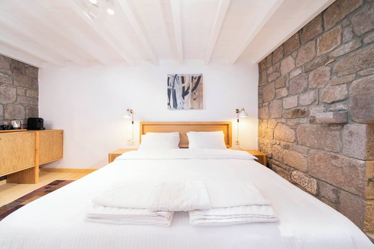A neatly made bed with white linens and towels, a wooden headboard, and a stone wall to the right.