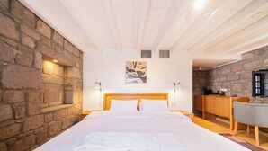 Une chambre d’hôtel avec un mur en pierre, une tête de lit en bois, un lit avec du linge de maison blanc, un tableau accroché au mur et un petit bureau avec une chaise.