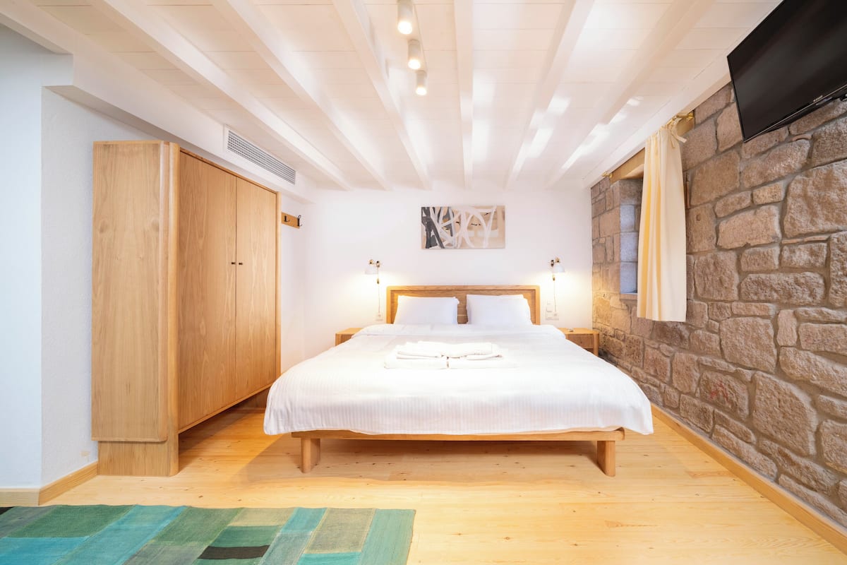 A bedroom with a wooden bed, a stone wall, a TV mounted on the wall, and a wardrobe.