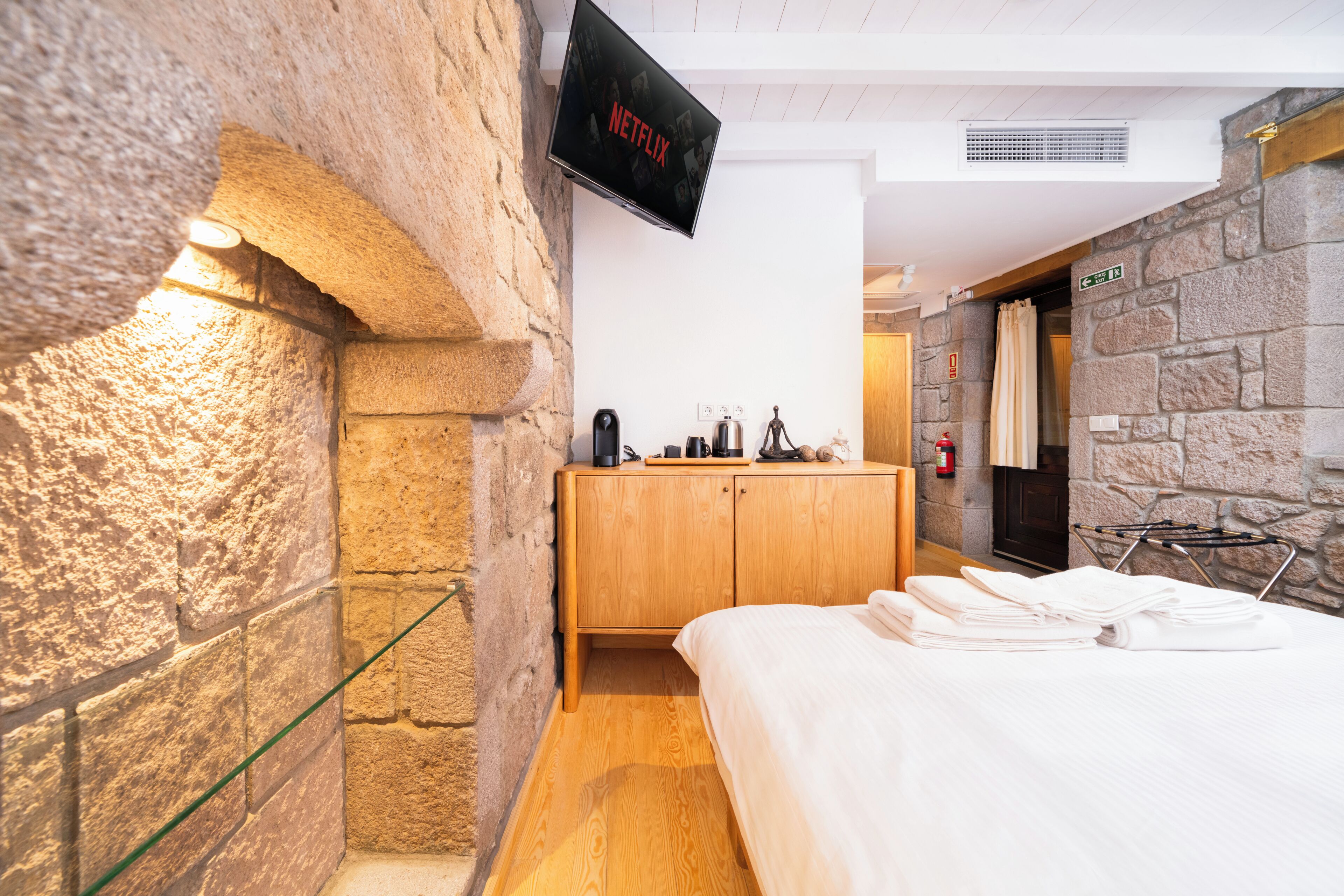 Une chambre d’hôtel avec un mur en pierre, un lit avec du linge de maison blanc, une armoire en bois, une machine à café et un téléviseur affichant le logo de Netflix.