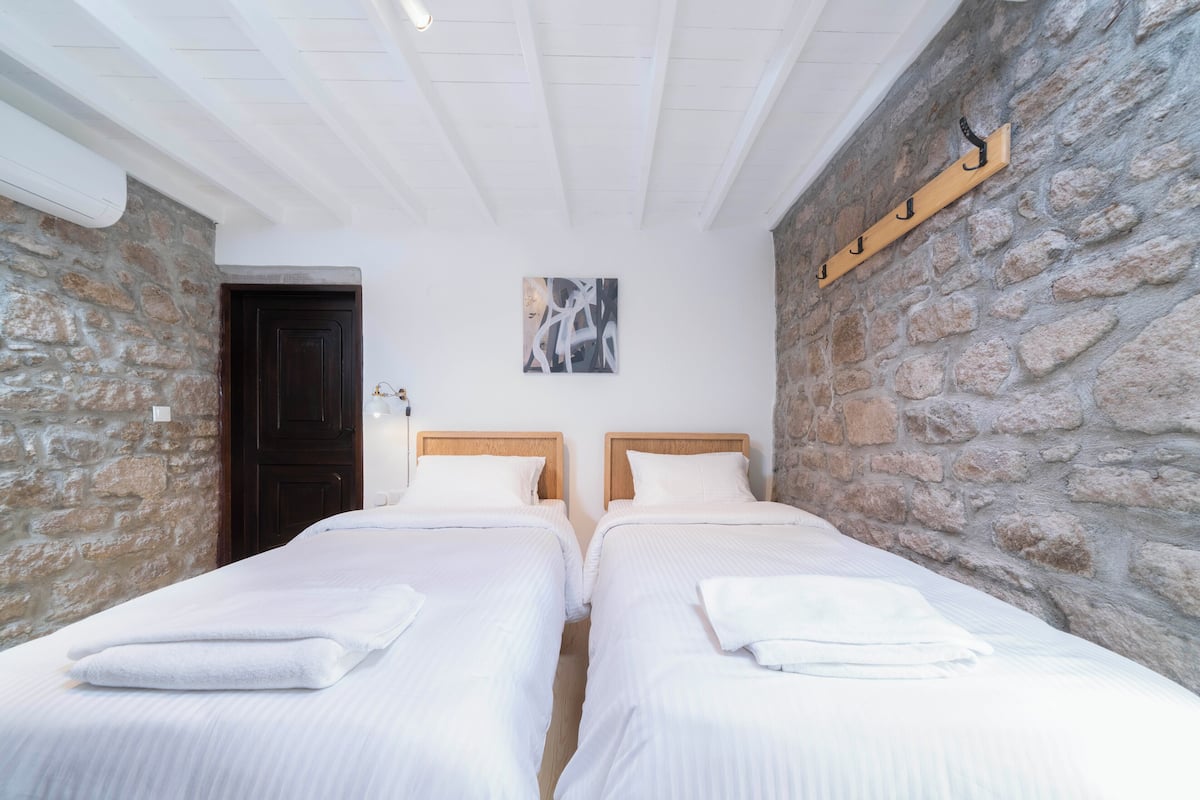 A room with two beds, white bedding, a stone wall, a wooden door, and a mounted coat rack.