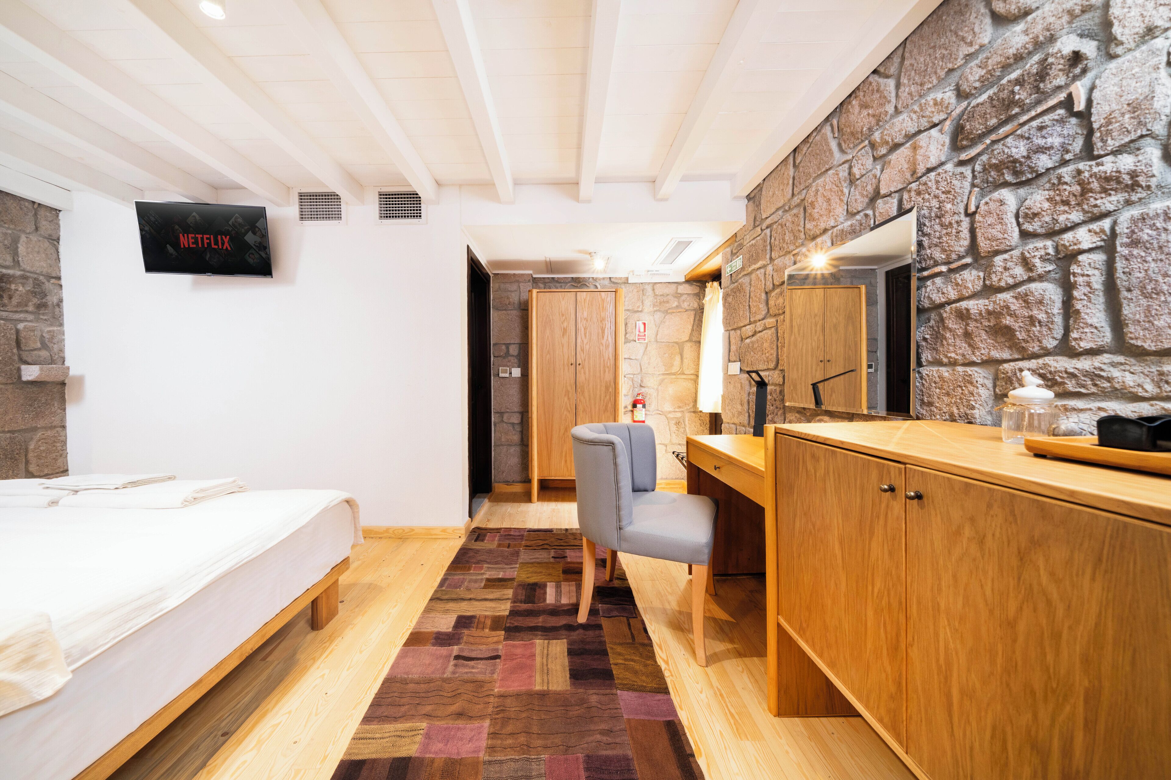 A bedroom with a bed, a desk, a chair, a TV, and a stone wall.