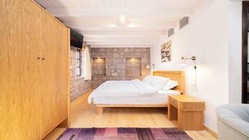 A bedroom with a wooden bed, a nightstand, a desk, a stone wall, and a television on the wall.