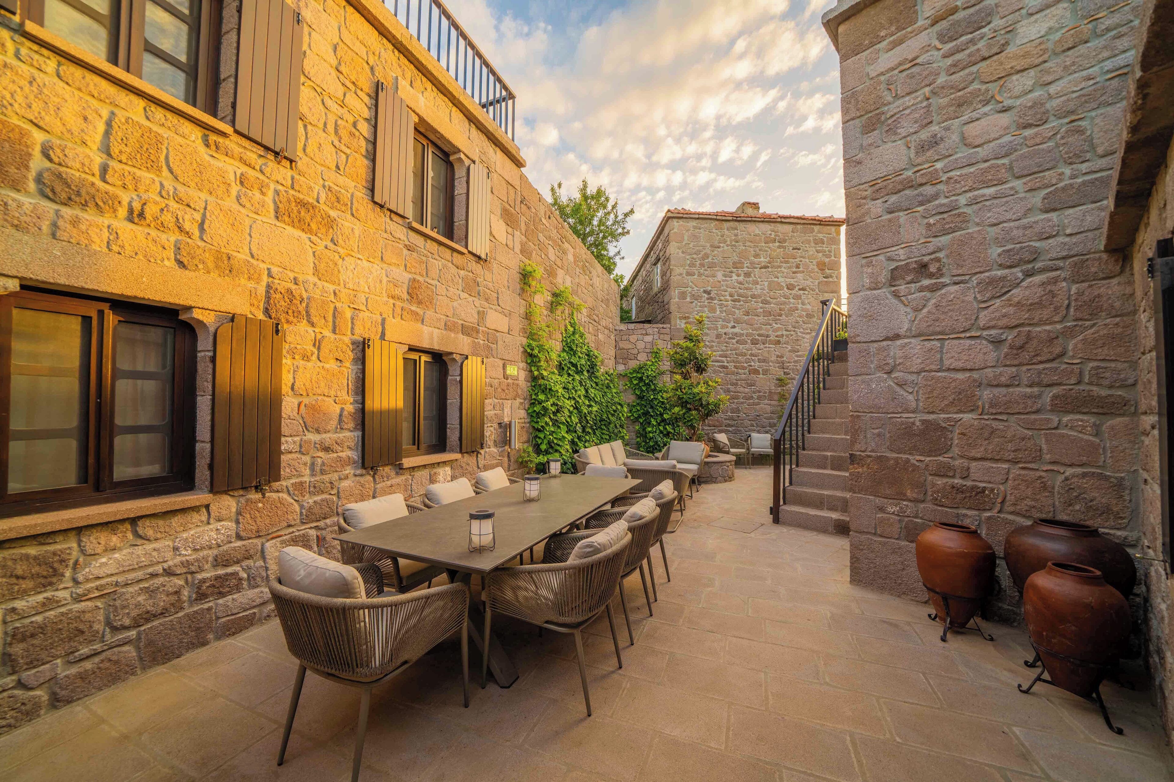 A stone building with a patio area featuring a long table and chairs, a staircase, and potted plants.