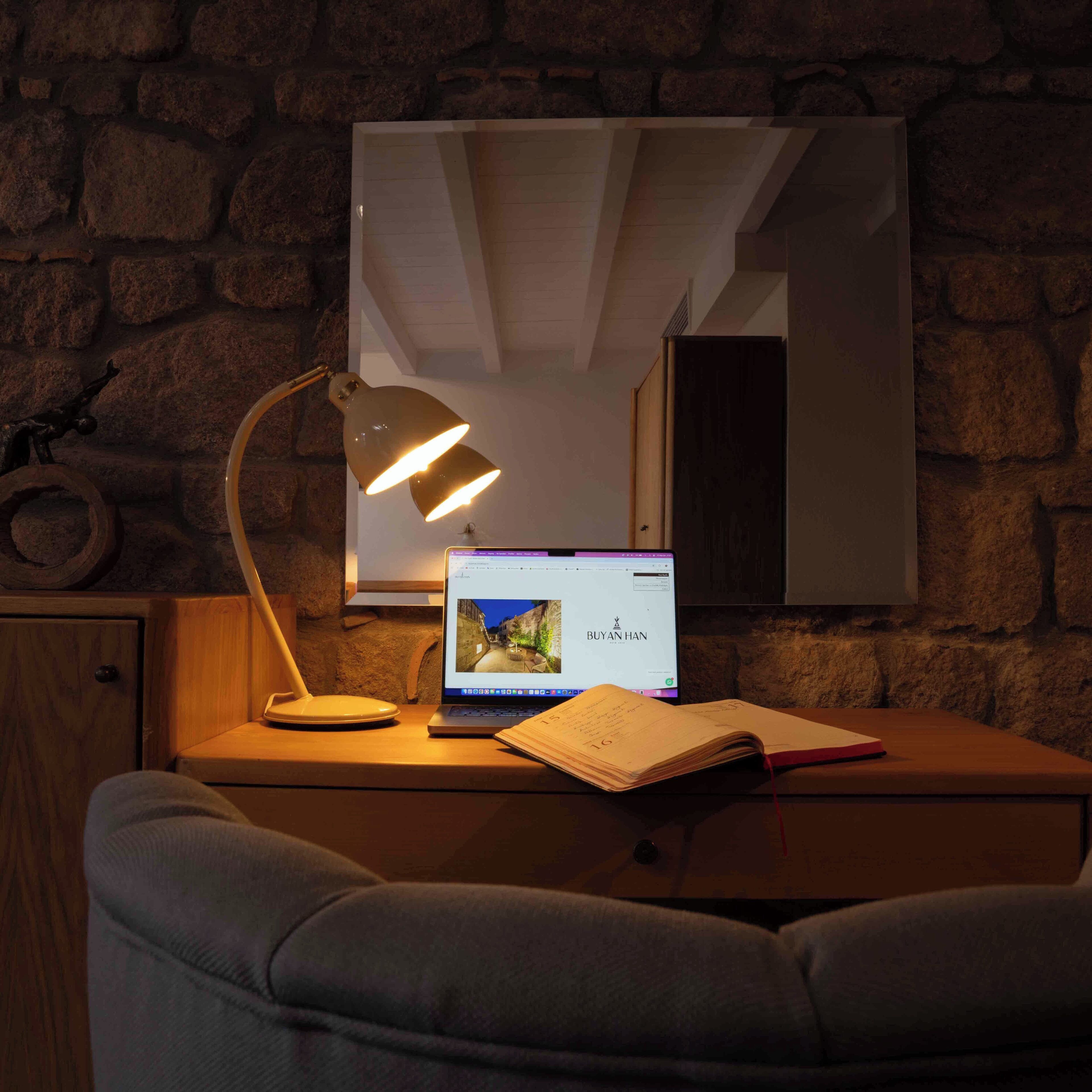 A study area with a laptop, a desk lamp, and a notebook on a wooden desk against a stone wall.